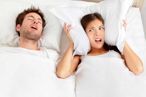 Man snoring while woman covers her ears with a pillow.